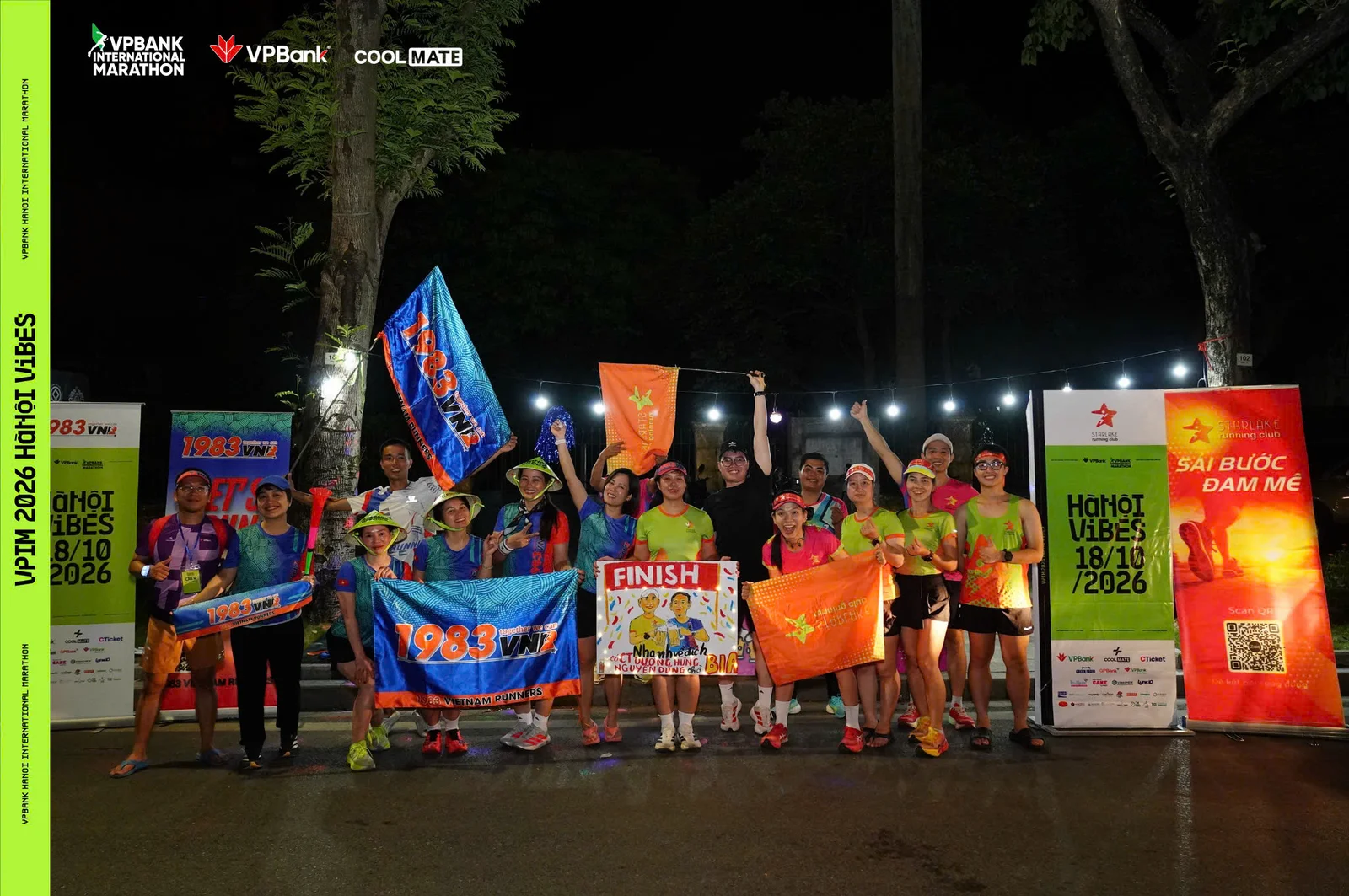 Đội cổ vũ Starlake cười tươi bên cờ cam, biển FINISH, banner Starlake Running Club và biển Hà Nội Vibes 18/10/2026 trên đường chạy VPIM 2026