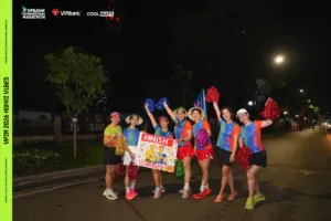 Nhóm cổ vũ giơ cao pom-pom đỏ, xanh, vàng bên tấm biển FINISH, phía xa là con đường vắng và những ngọn đèn đêm