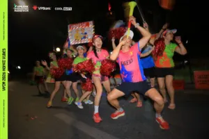 Một thành viên nửa ngồi nửa đứng thổi kèn vuvuzela giữa đội cổ vũ cầm pom-pom đỏ trong đêm
