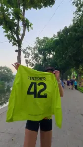 Một chị giơ áo FINISHER 42km khoe sau khi về đích, phía sau là mặt hồ và hàng cây buổi sớm